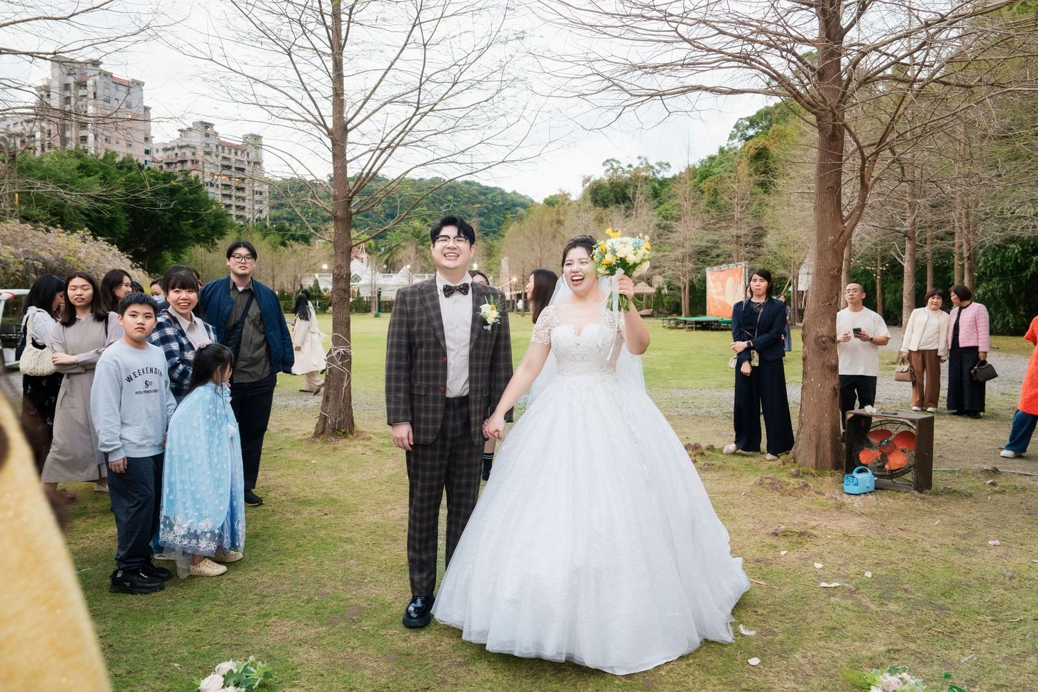 Wedding party gathers for outdoor photos in a natural park setting with trees and grass on a sunny day.