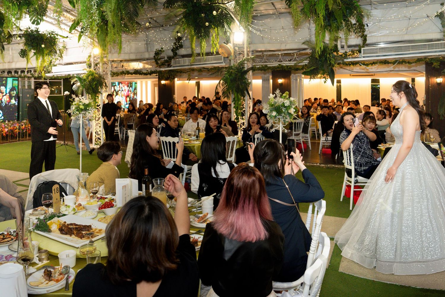 An elegant outdoor wedding reception with guests seated at tables under hanging greenery and string lights.