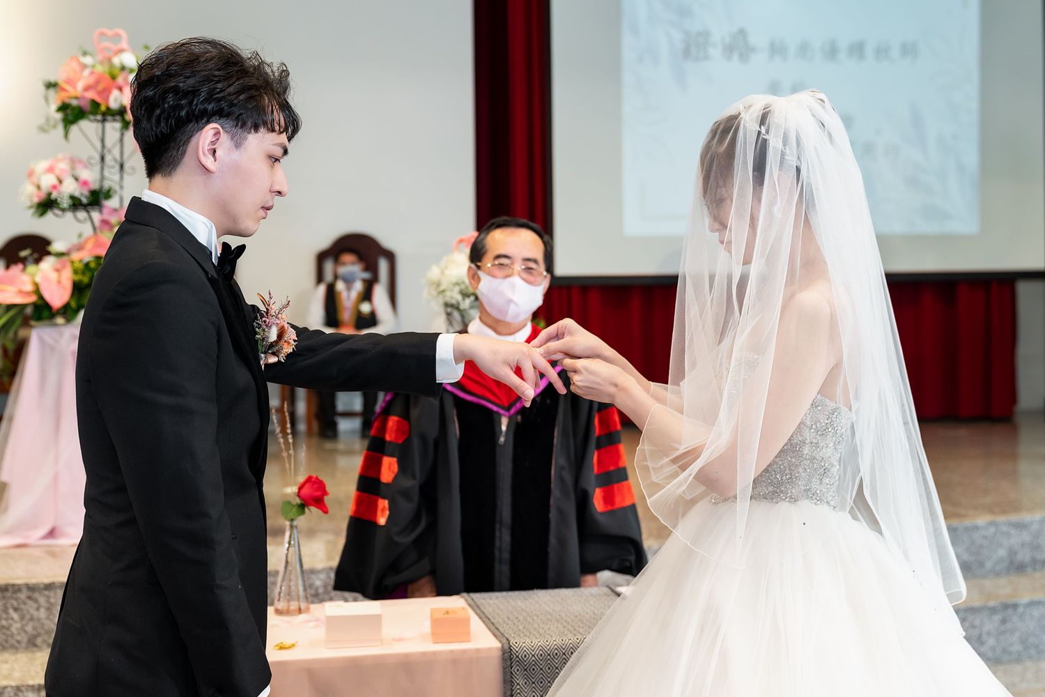 A wedding ceremony with the couple exchanging rings while an officiant in red robes observes.