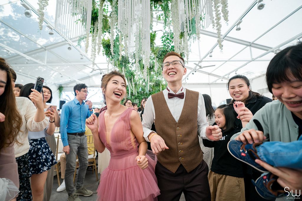 Wedding guests celebrating enthusiastically under hanging greenery in a bright indoor venue.