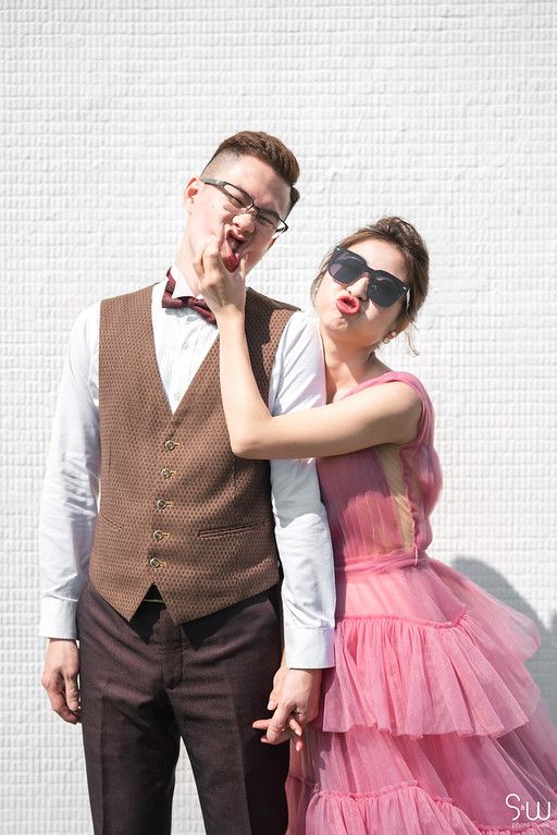 A stylish pair pose together against a white wall wearing formal attire with a pink tulle dress and brown vest.