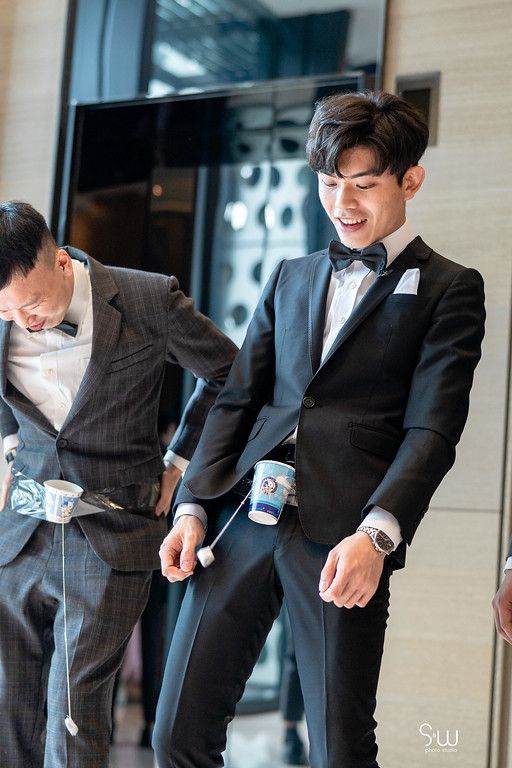 Two groomsmen in formal wear laugh and pose in a modern hotel hallway.