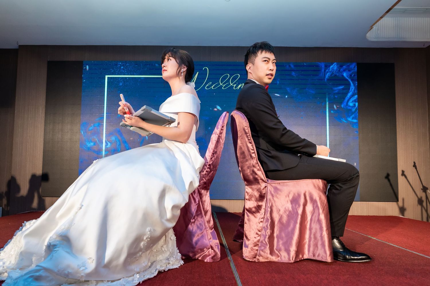Bride and groom sit back-to-back on pink chairs during wedding reception game.