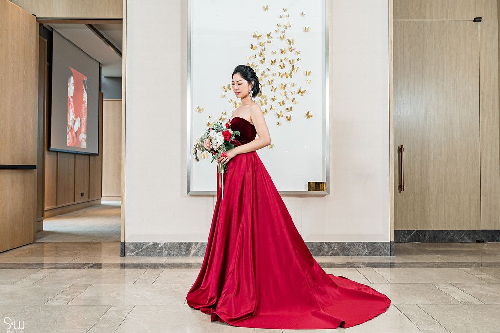 Bride in flowing red gown with long train poses elegantly against wall with golden butterfly art.