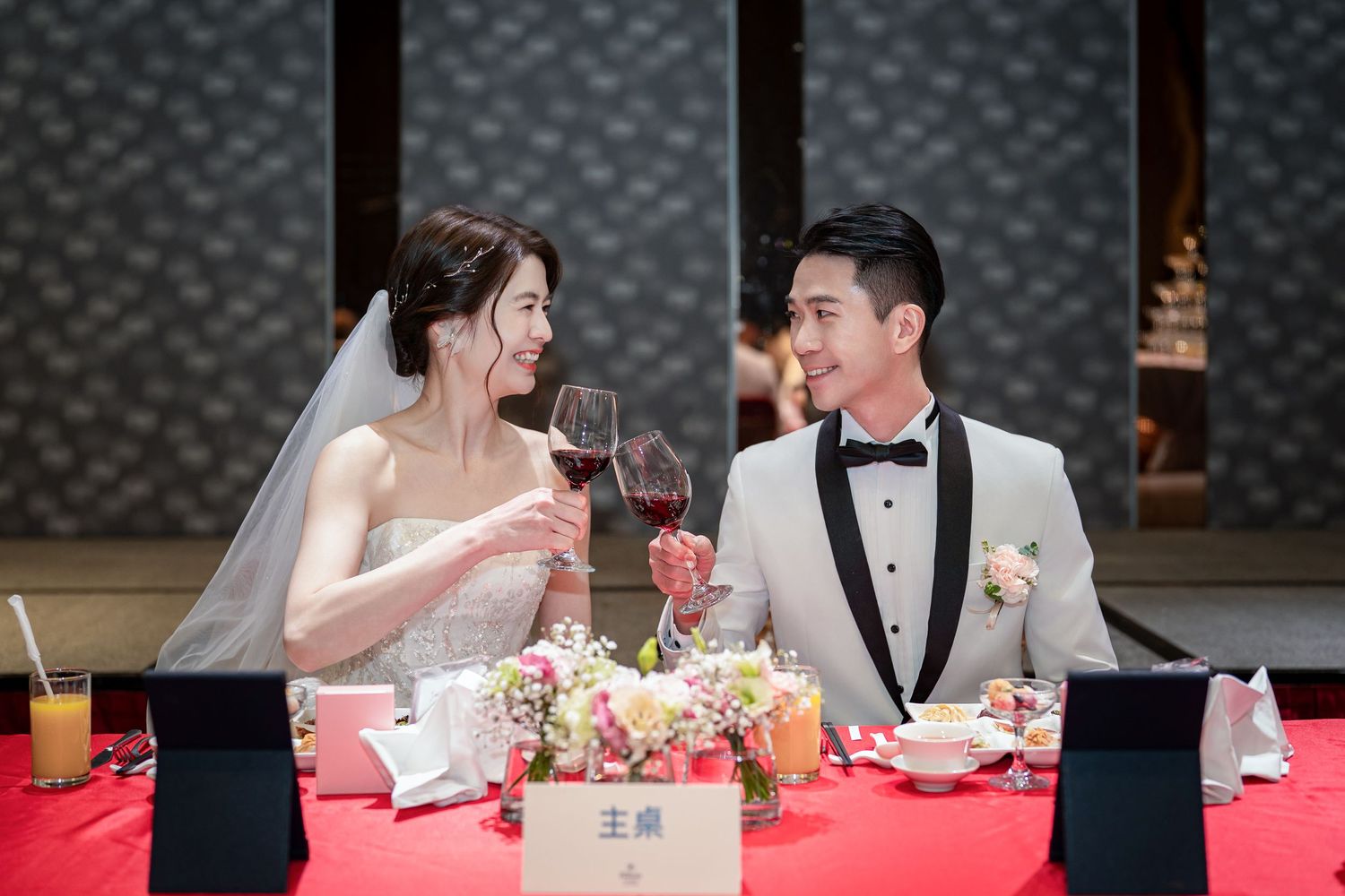 A bride and groom share wine glasses at their wedding reception table decorated with floral centerpieces.