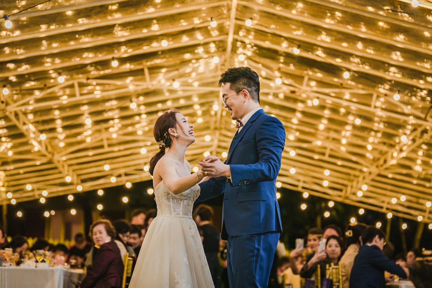 A couple shares their first dance at an outdoor wedding reception under warm string lights at dusk.