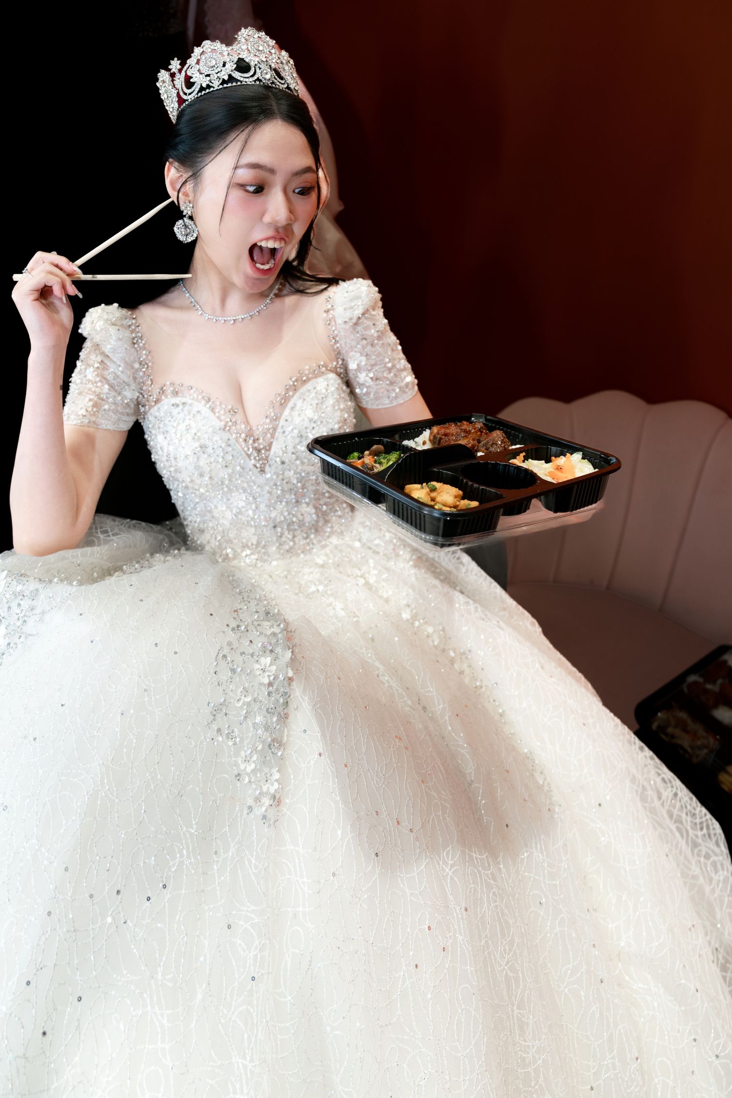 A bride in a sparkly white ball gown and tiara shows excitement while looking at food on a tray.