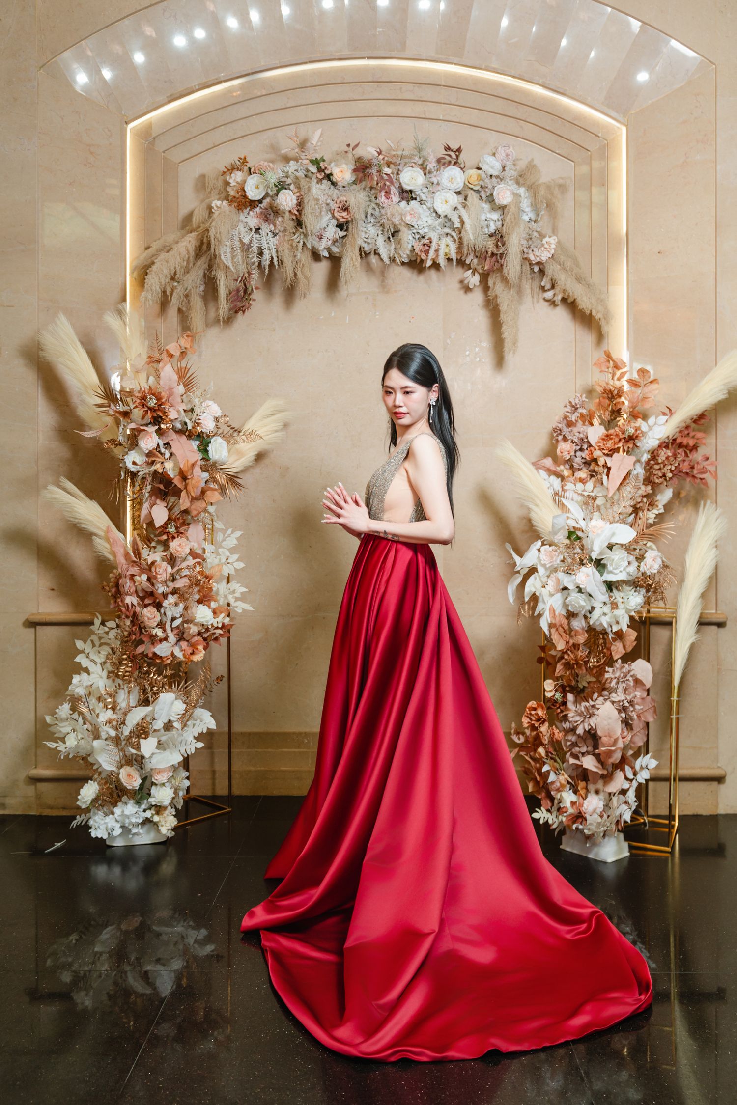A person in a flowing red satin gown poses under an elegant floral arch with dried pampas grass and white blooms.