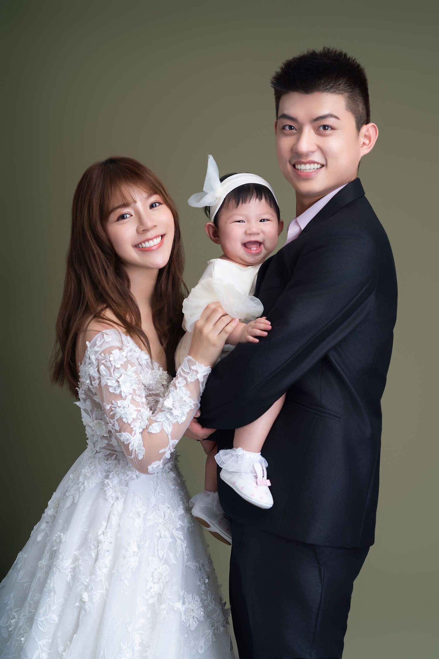 A family portrait of parents in formal attire holding their smiling baby wearing white against a dark background.
