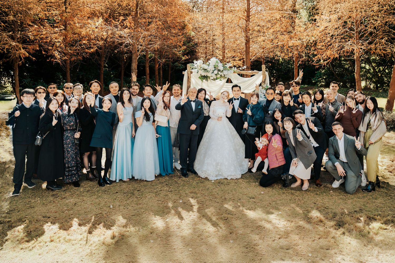 A large wedding party poses together outdoors on an autumn day with golden fall foliage in the background.