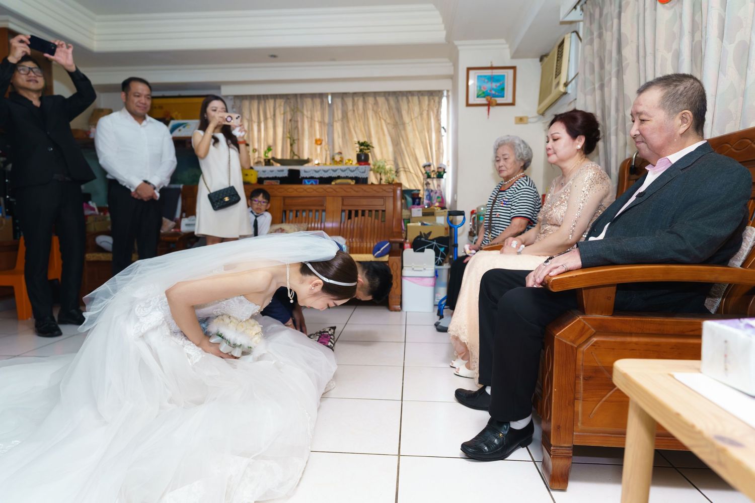 Wedding ceremony attendees seated in a living room watch as someone in a white wedding dress bows down to the floor.