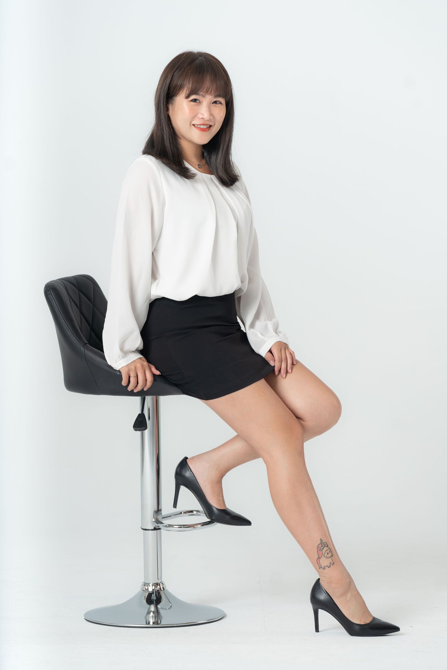 A model in a white sweater and black shorts poses elegantly on a modern chrome bar stool against a white backdrop.