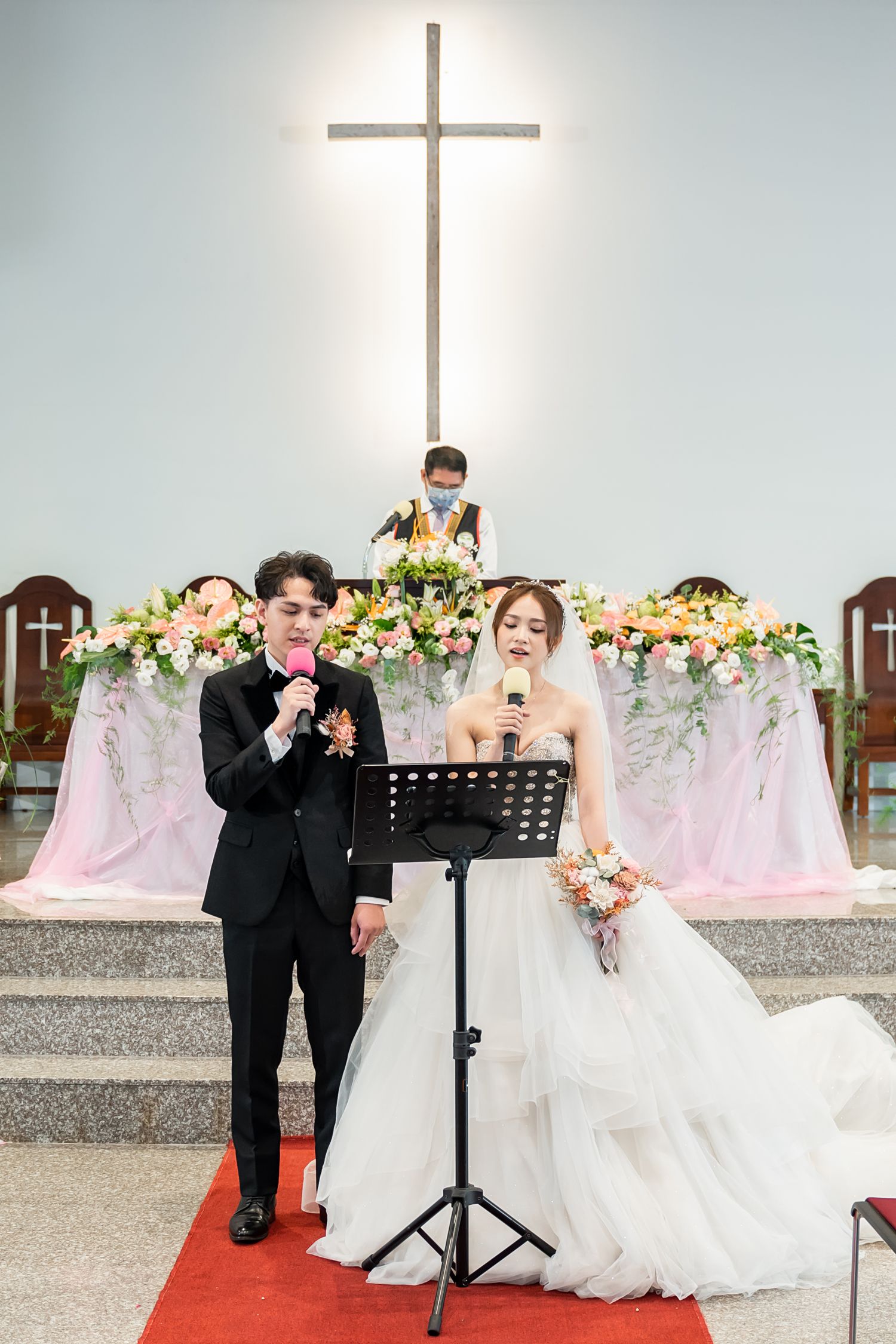 Wedding couple performing at altar beneath illuminated cross with floral decorations.