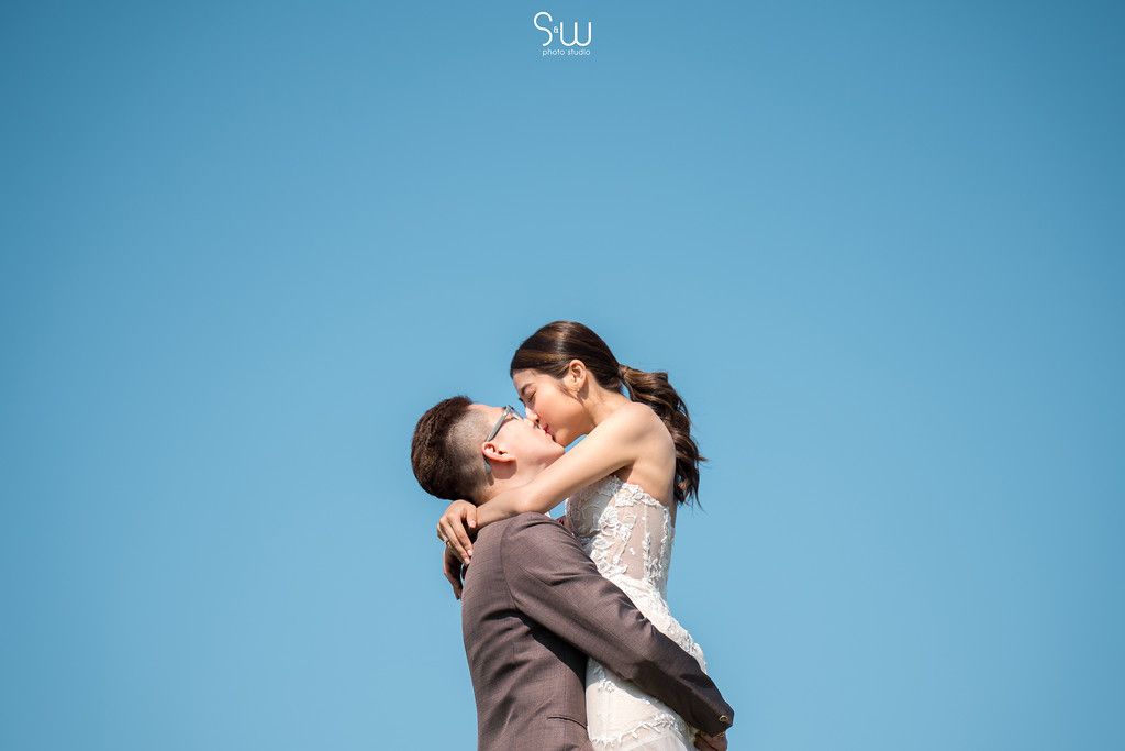 Romantic wedding couple embrace against bright blue sky.