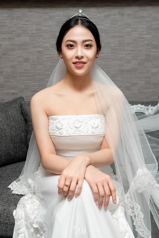 A bride wearing a classic white wedding dress and veil sits poised on a grey couch.