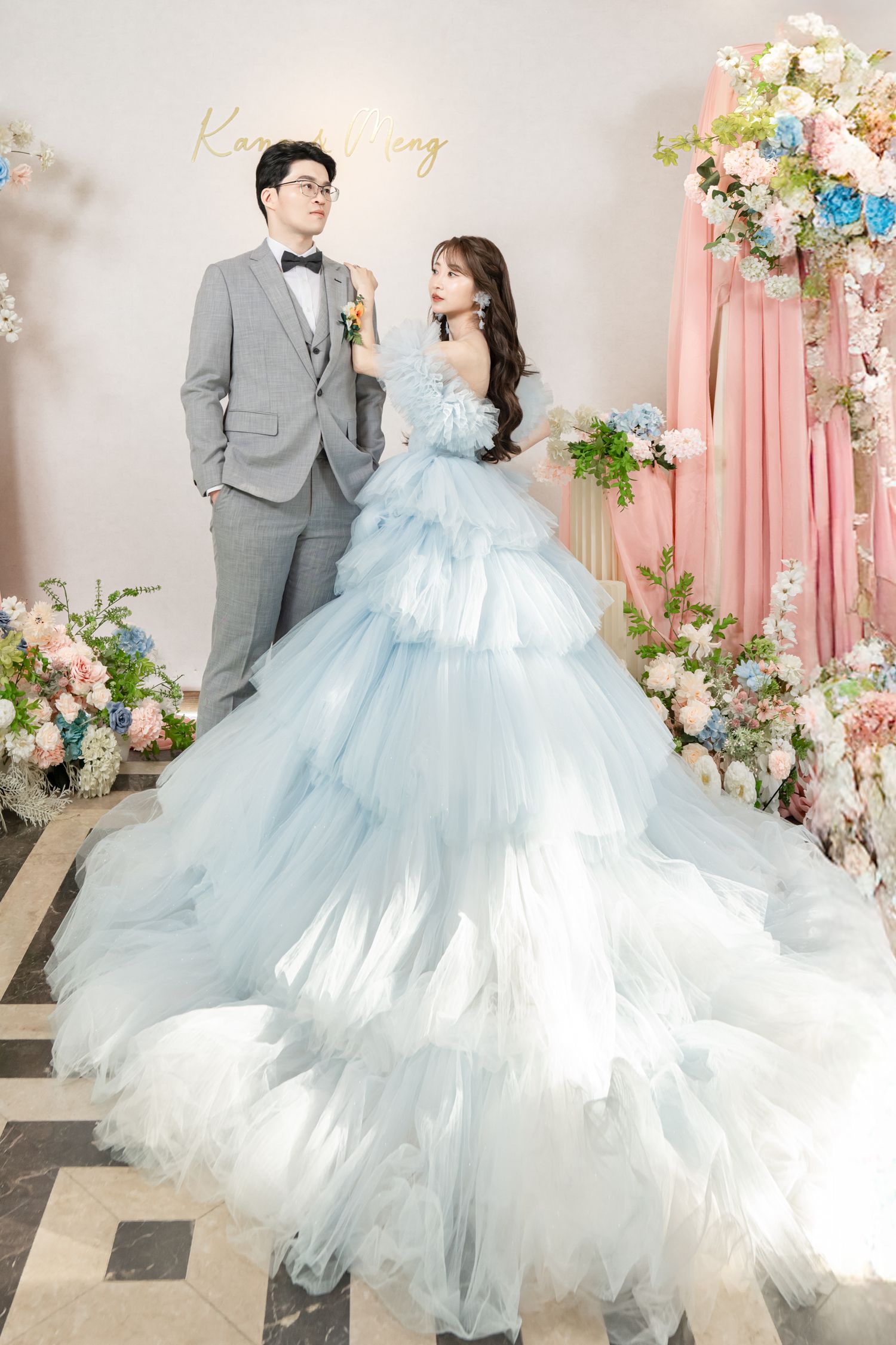 A couple poses in wedding attire with the bride wearing a light blue tulle gown against a floral backdrop.