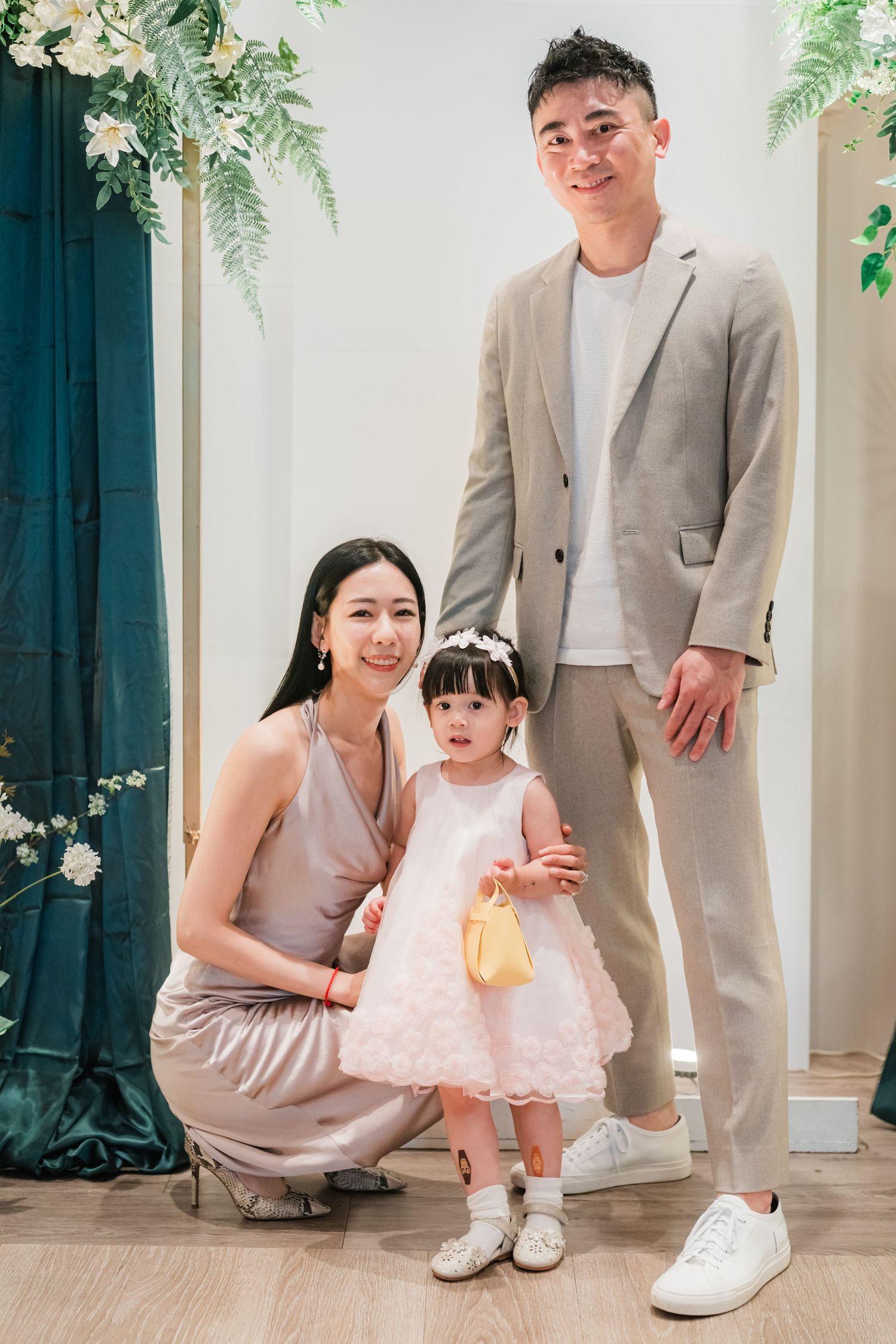 Adult in beige suit and sneakers poses with child in pink dress against light background.