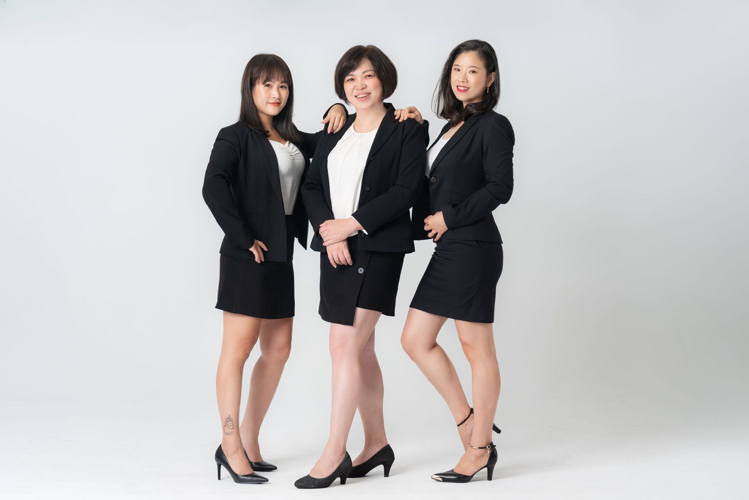 Three women in black outfits pose together in a studio setting with white background.