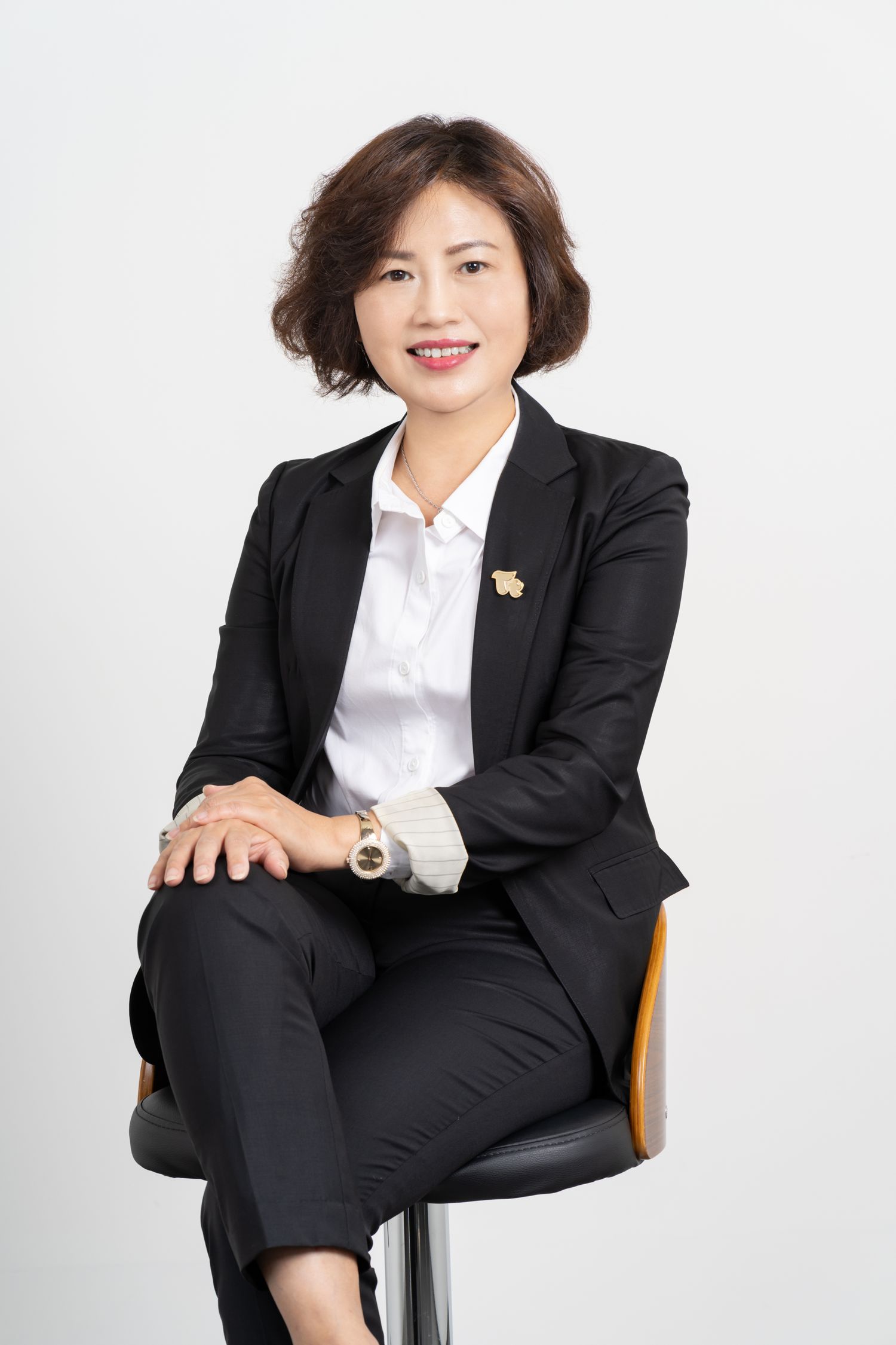 Professional business portrait of someone wearing a black suit and white shirt seated on a chair against light background.