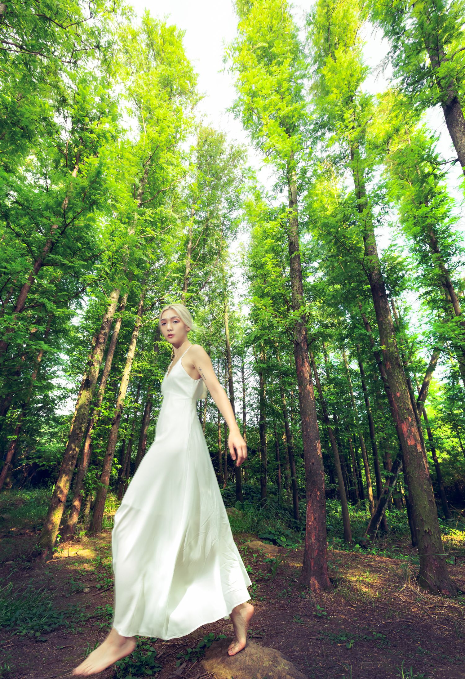A graceful figure in a flowing white dress twirls barefoot in a sunlit forest setting.
