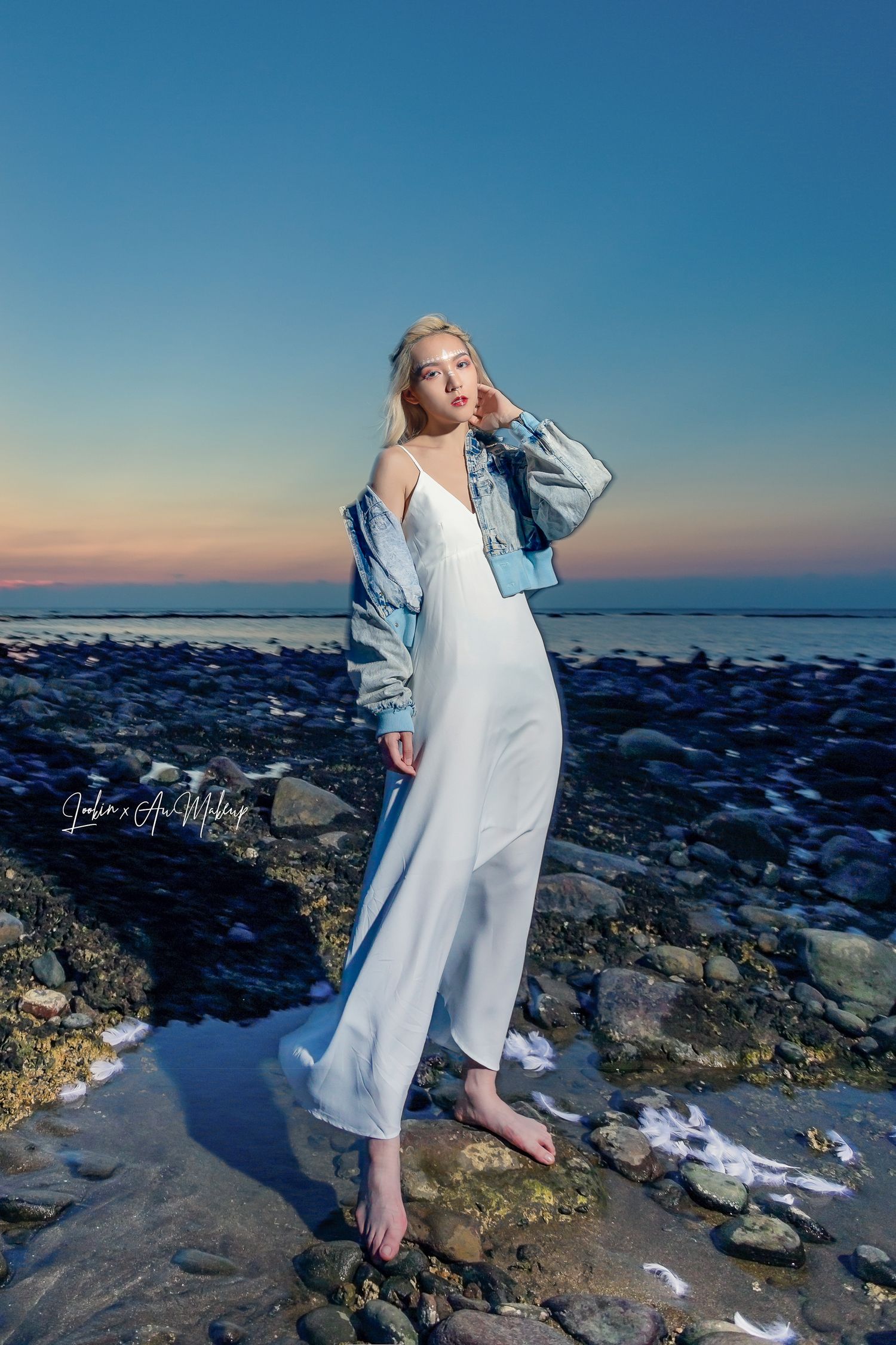A person in a flowy white dress and denim jacket poses on rocky coastal terrain at sunset against a blue sky.