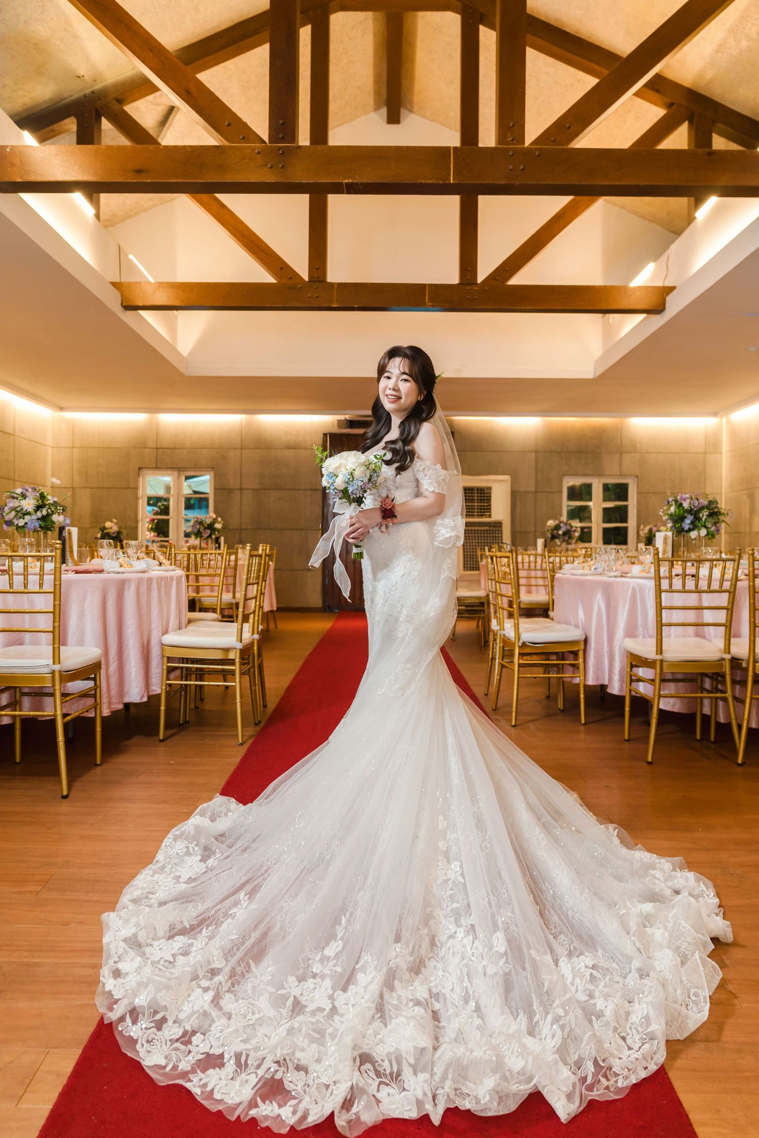 A bride in a stunning lace mermaid wedding dress with long train stands in an elegant reception venue with wooden beams.