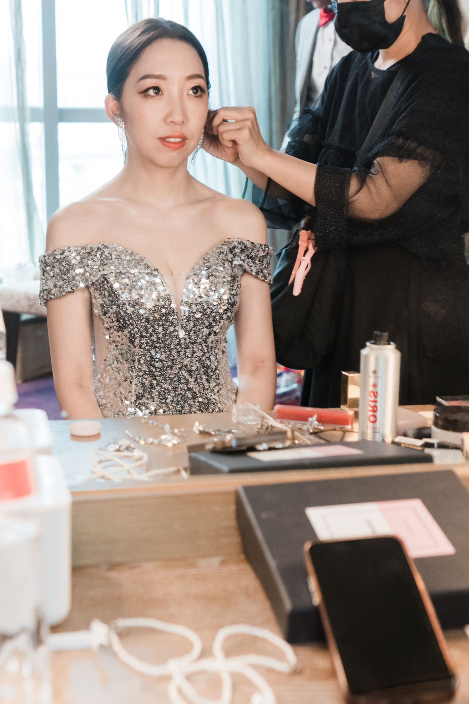 A makeup artist works on a client wearing a sparkly silver off-shoulder gown while getting ready at a vanity table.