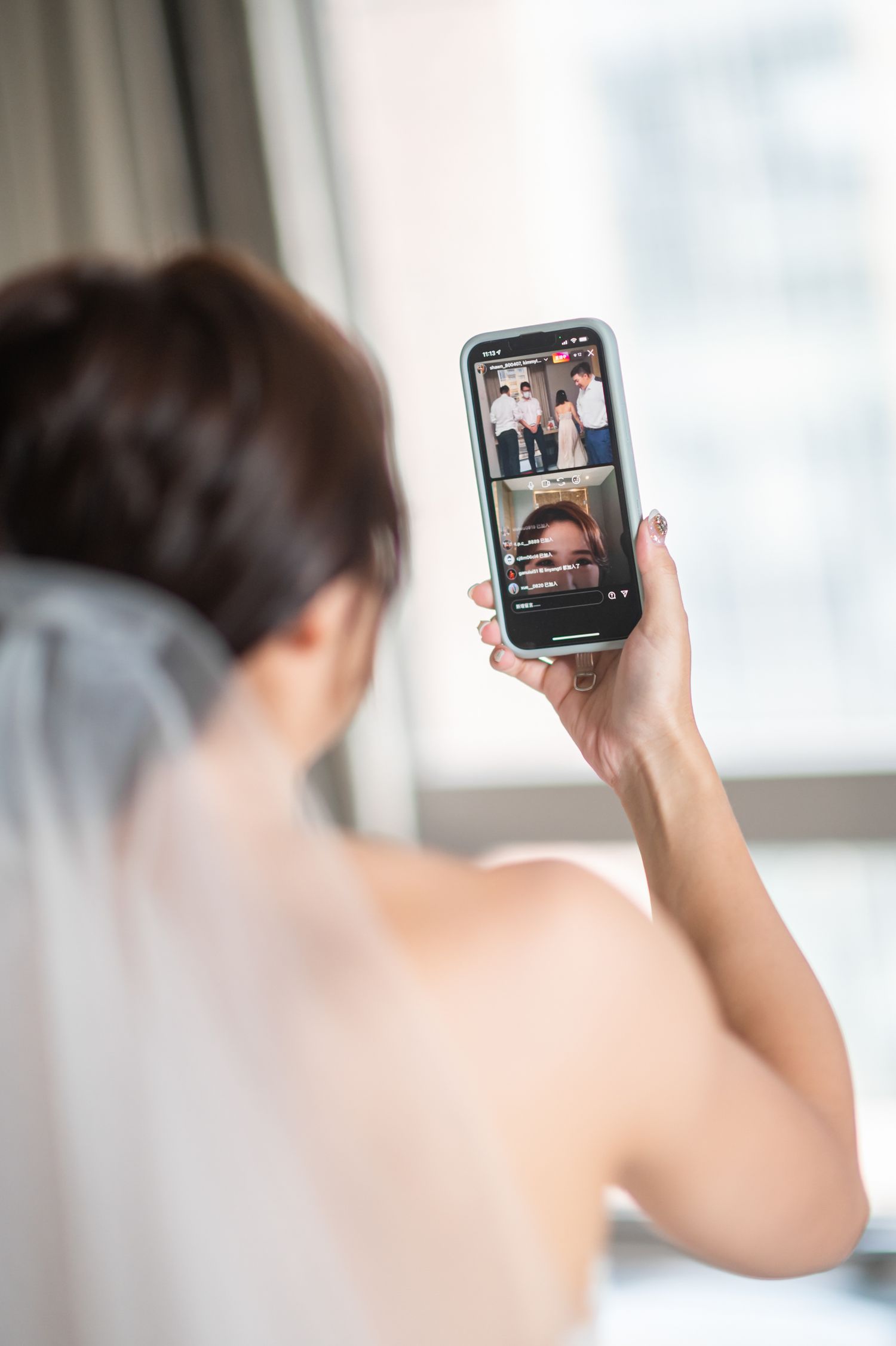 Someone holds a smartphone showing a video chat or social media app while sitting near a window.