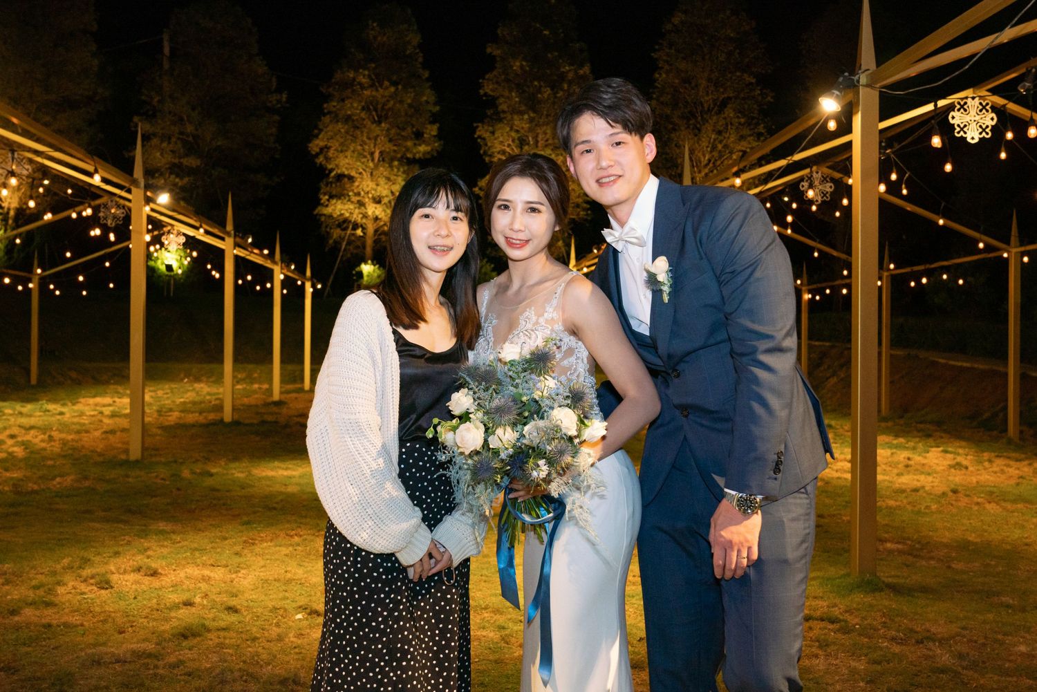 Three people pose together at an outdoor evening wedding reception under string lights with a floral bouquet.