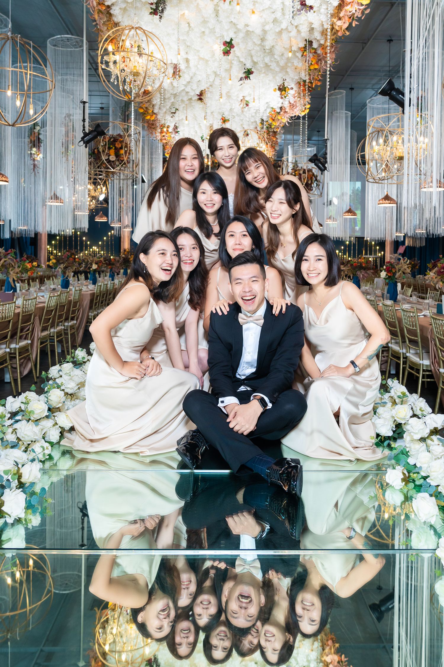 Glamorous wedding party poses on reflective floor under crystal chandeliers and floral decorations at luxury venue.