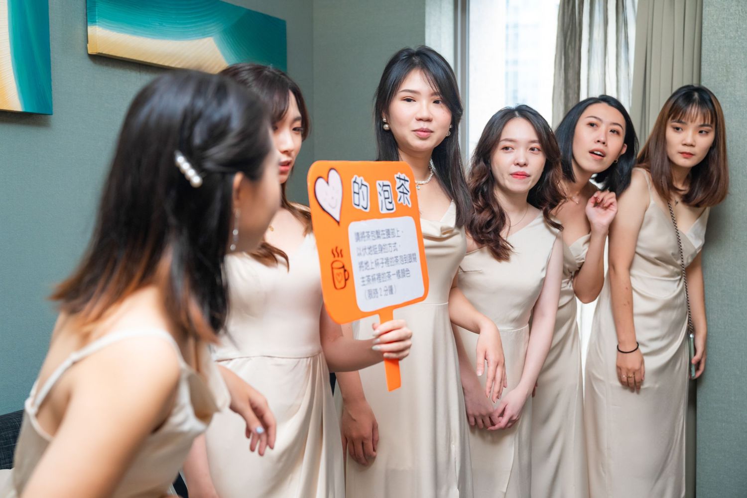 Bridesmaids in matching beige dresses pose playfully in a line while holding a wedding sign during preparations.