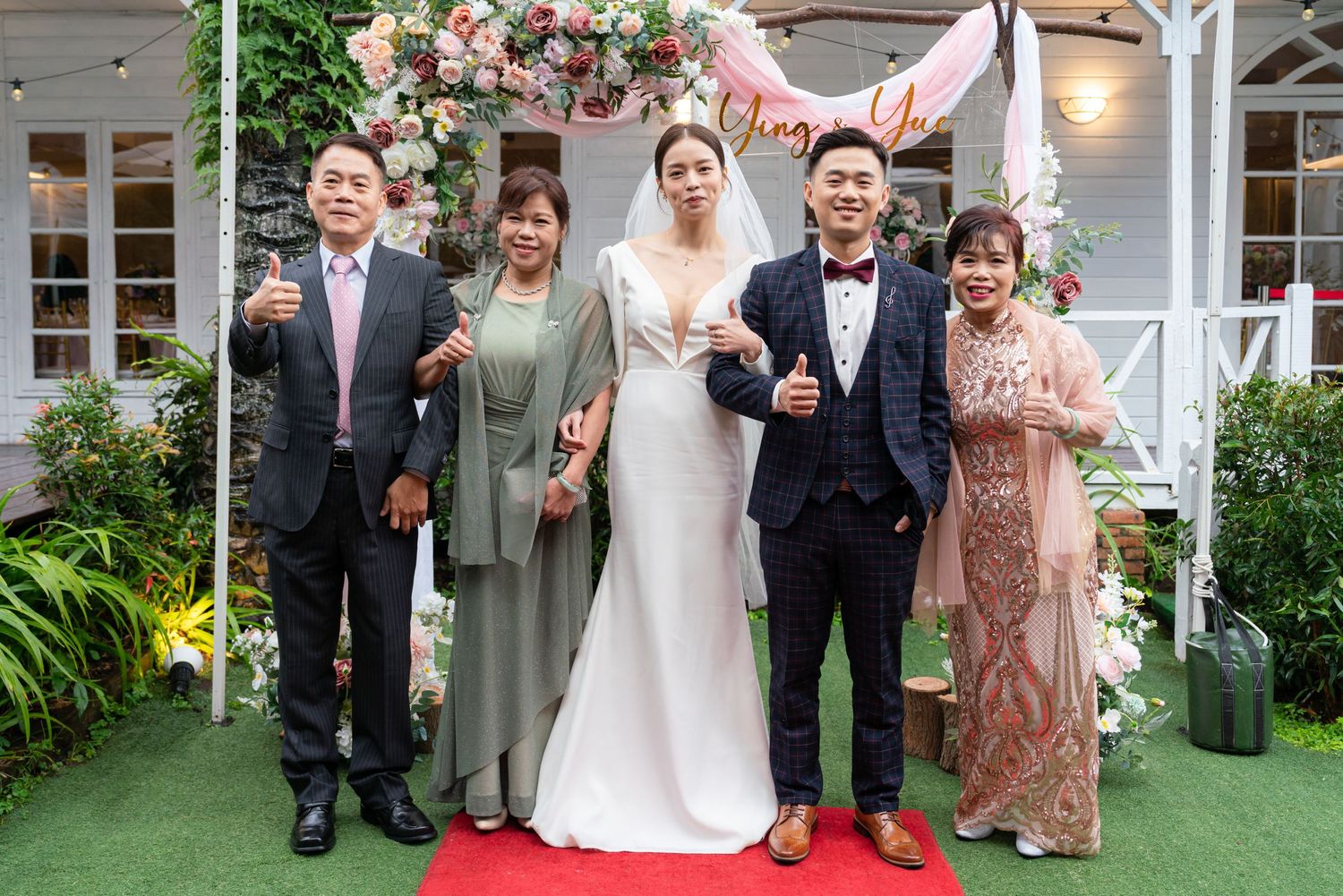 A wedding party stands together on a red carpet in front of a white gazebo with green foliage in the background.