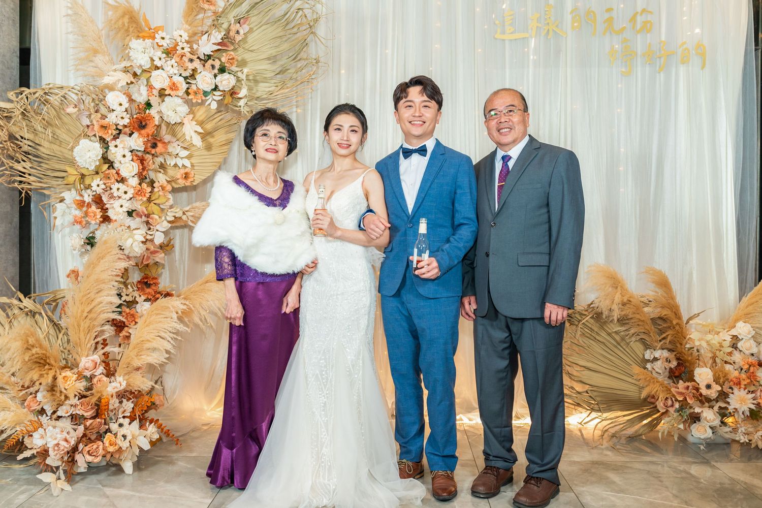 Four people pose in formal attire against an elegant backdrop with autumn-colored floral decorations at a wedding celebration.