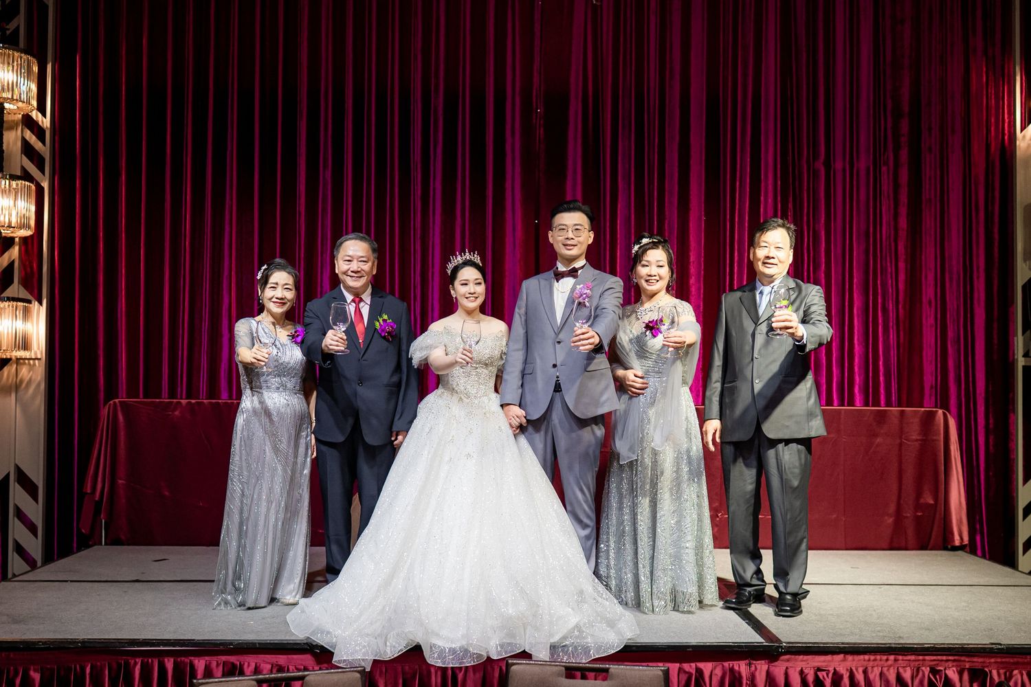 Members of a wedding party stand on stage with red velvet theater curtains in the background during a special event.