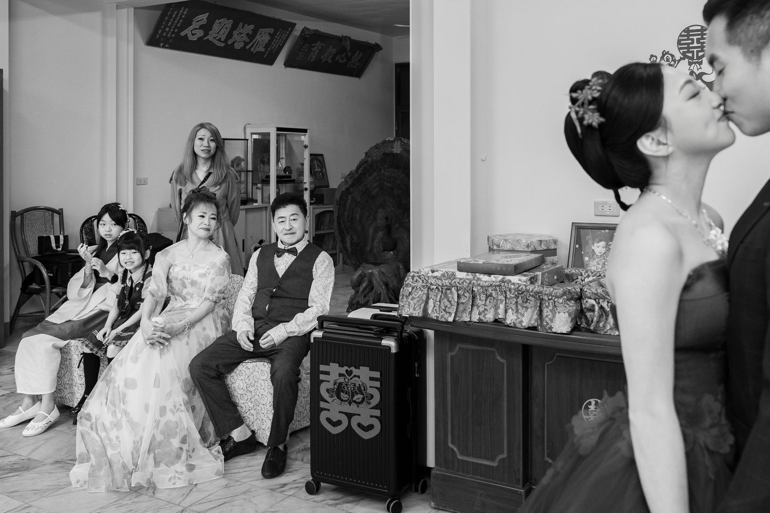 Black and white wedding photo showing couple kissing while family watches during traditional Chinese wedding ceremony.