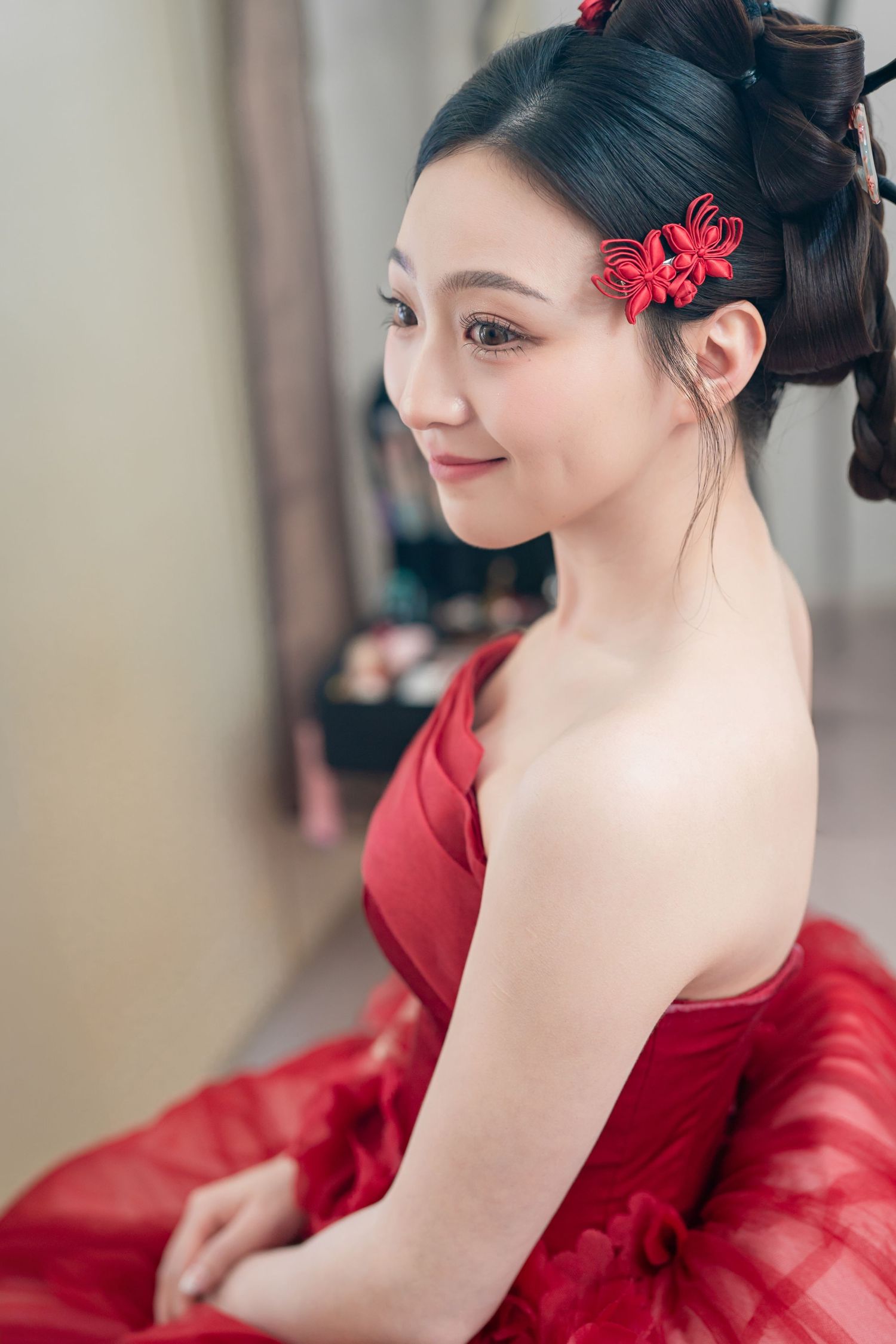 Young Asian woman in red strapless gown with red floral hair accessories, smiling gracefully in a bridal setting.