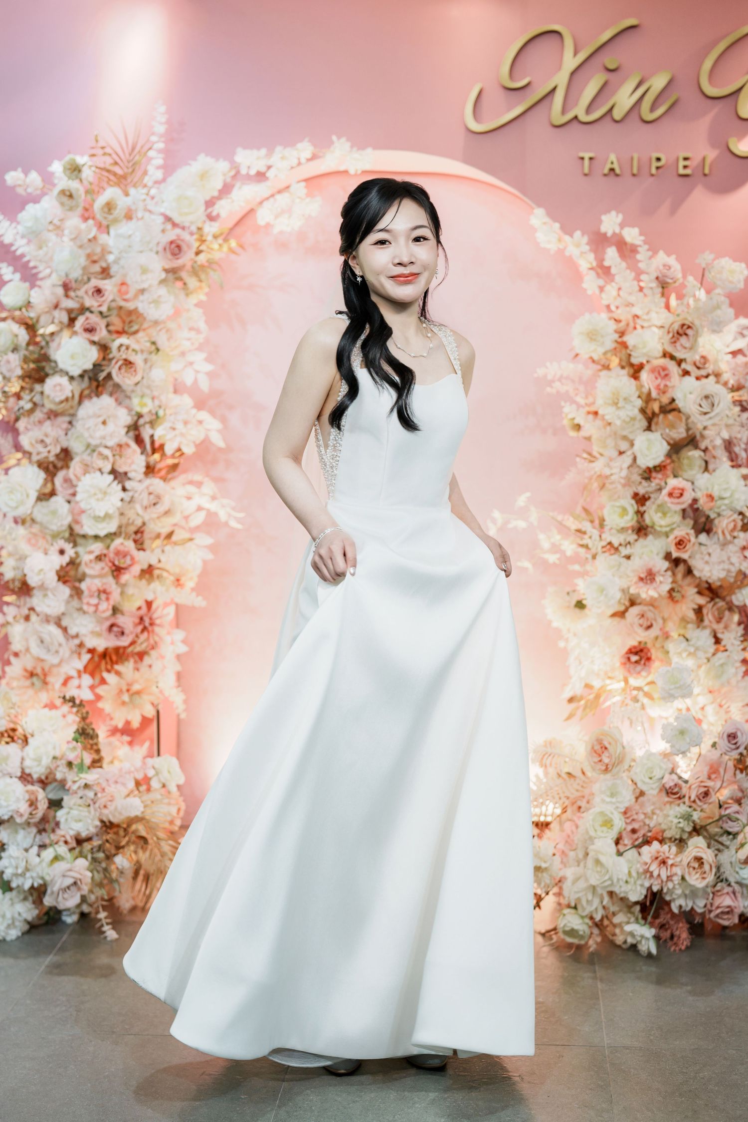 A bride in an elegant white wedding gown poses in front of a romantic floral arch backdrop with pink and peach blooms.