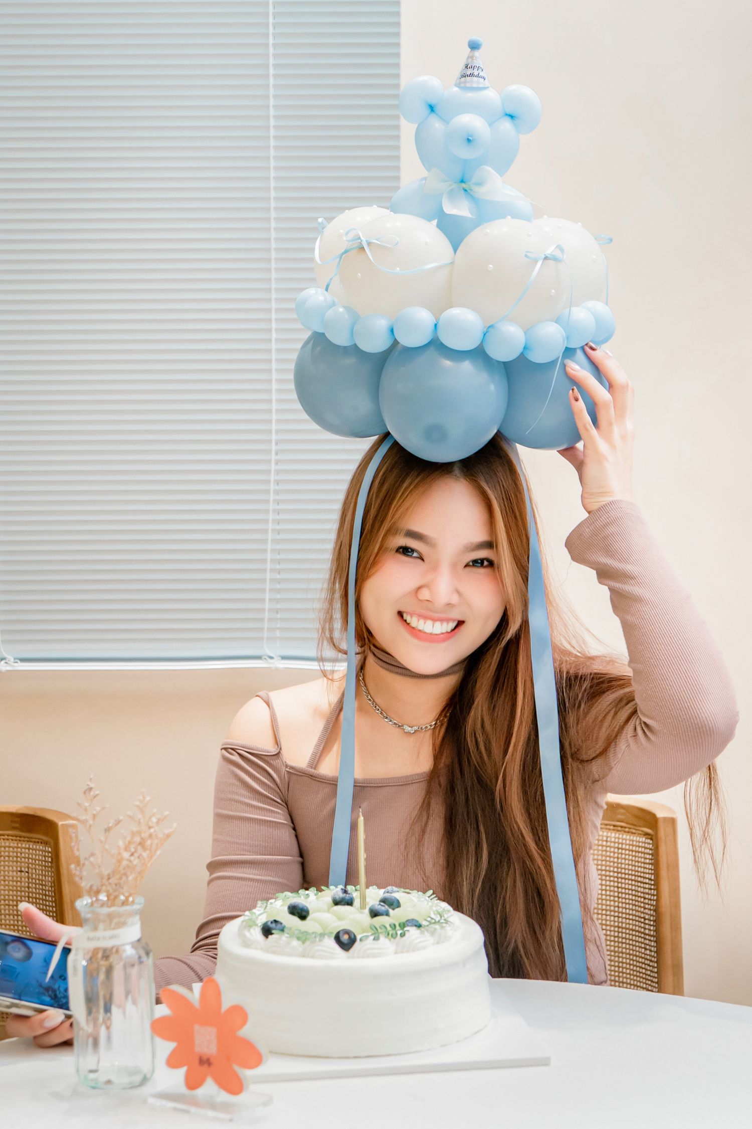 A birthday celebration setup with a white cake and blue balloon decorations featuring a teddy bear topper.