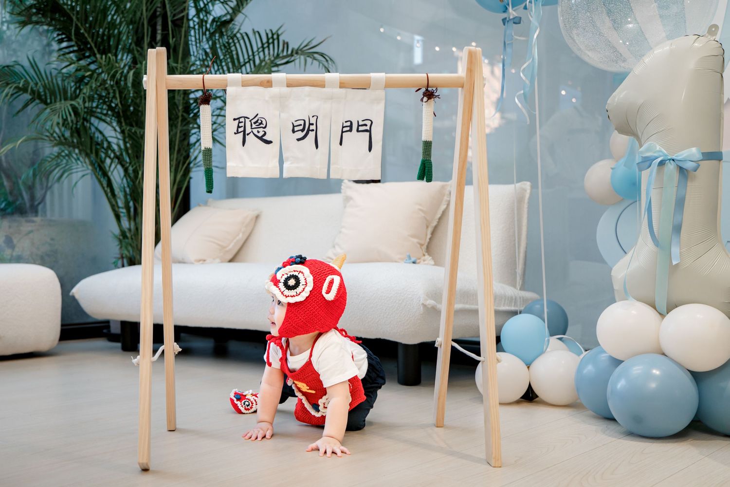 A baby crawls under a white table while surrounded by soft blue and white balloon decorations at a party.