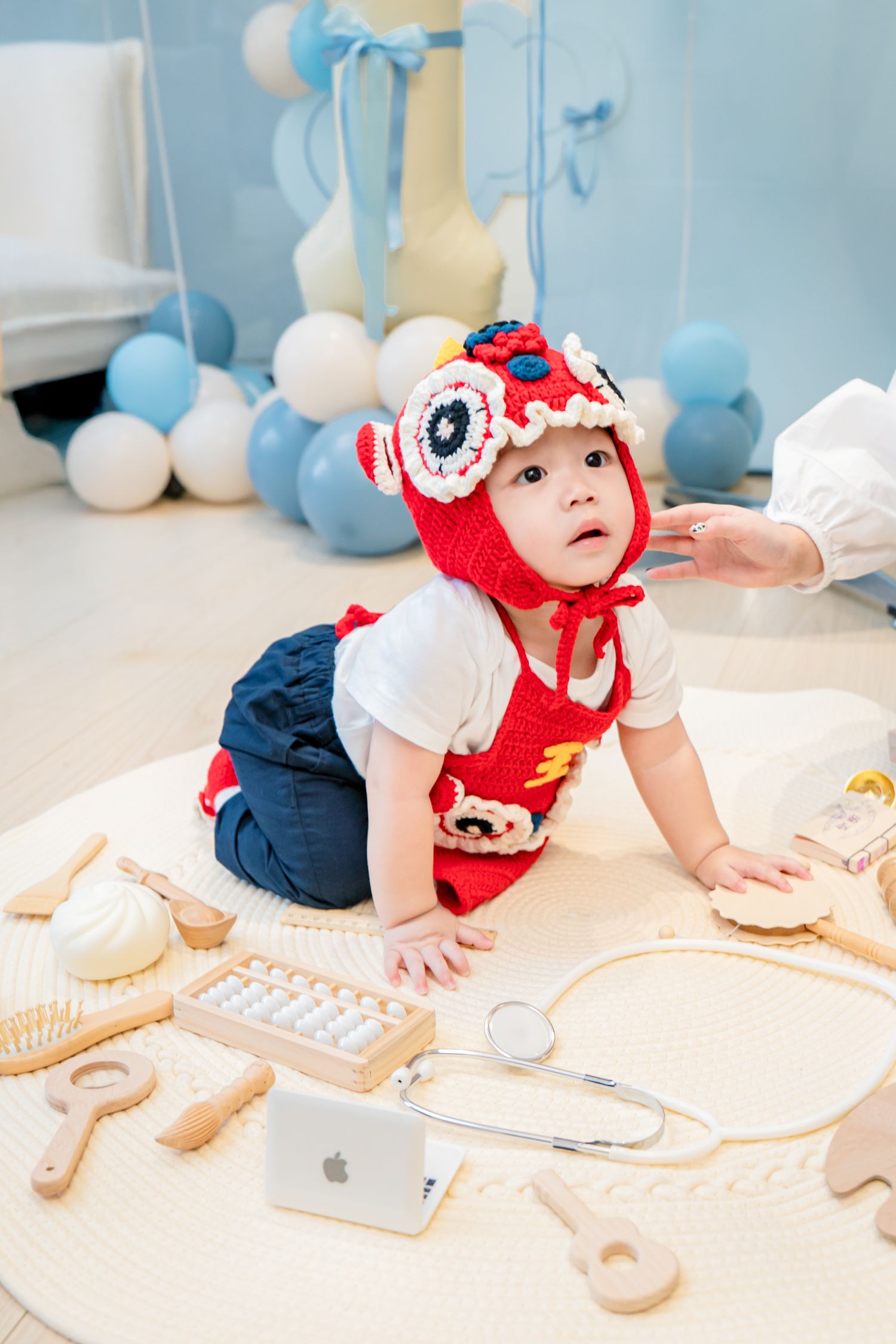 Small child in red knit hat and overalls crawls on white surface with toy medical instruments and balloons in background.