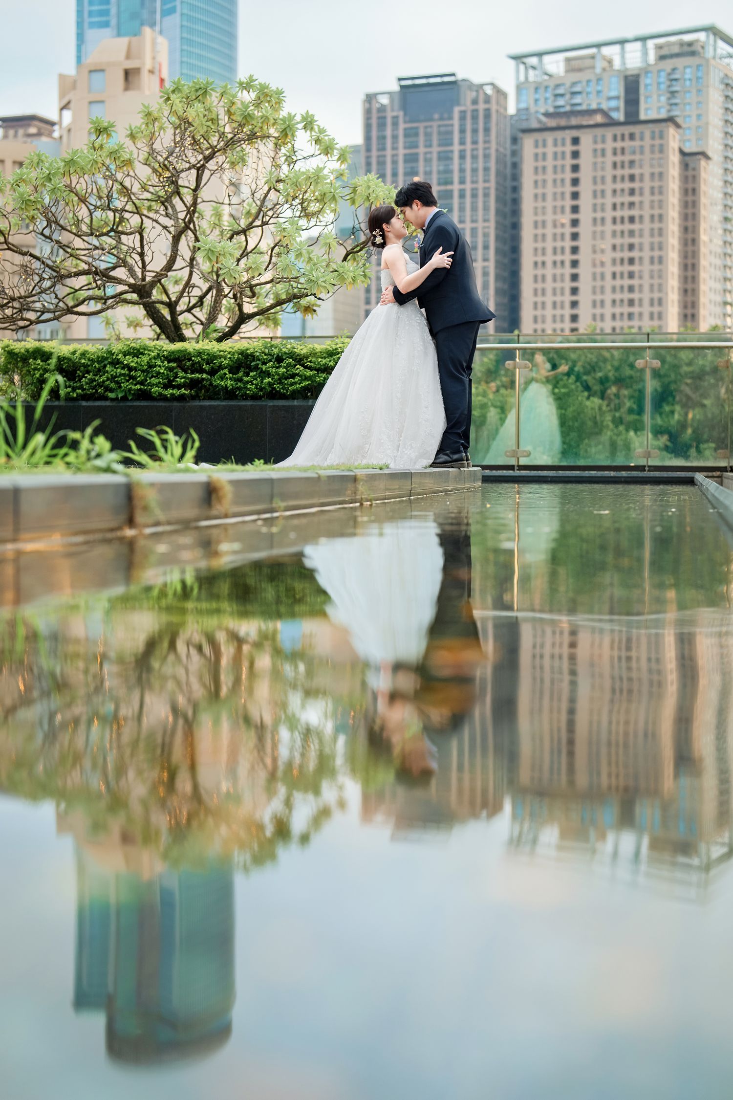 Wedding couple embraces by reflective pool with modern city skyline and skyscrapers in background.
