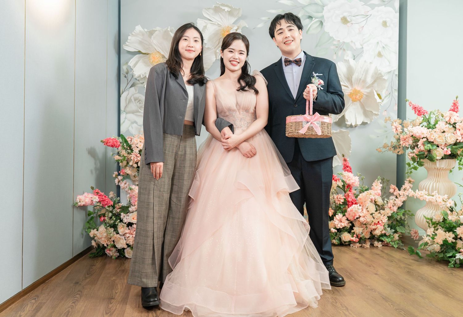 A wedding celebration with pink floral decorations and three people posing together against a light backdrop.