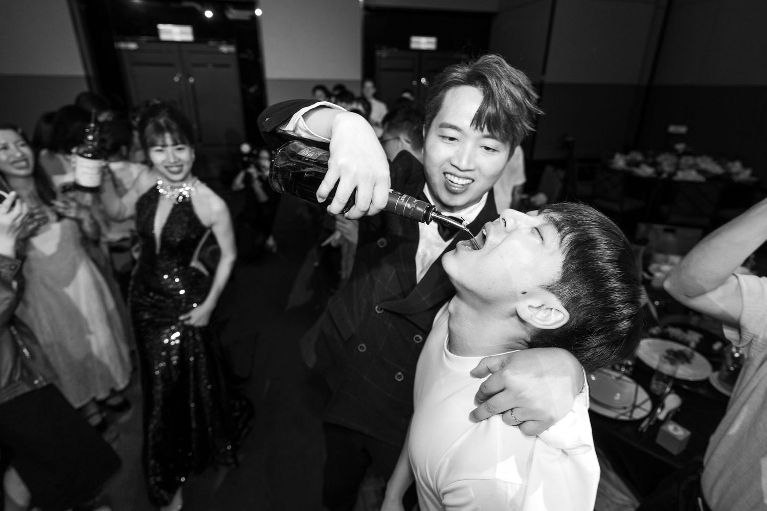 Black and white photo of guests celebrating at a wedding reception, with one person pouring champagne into another's mouth.
