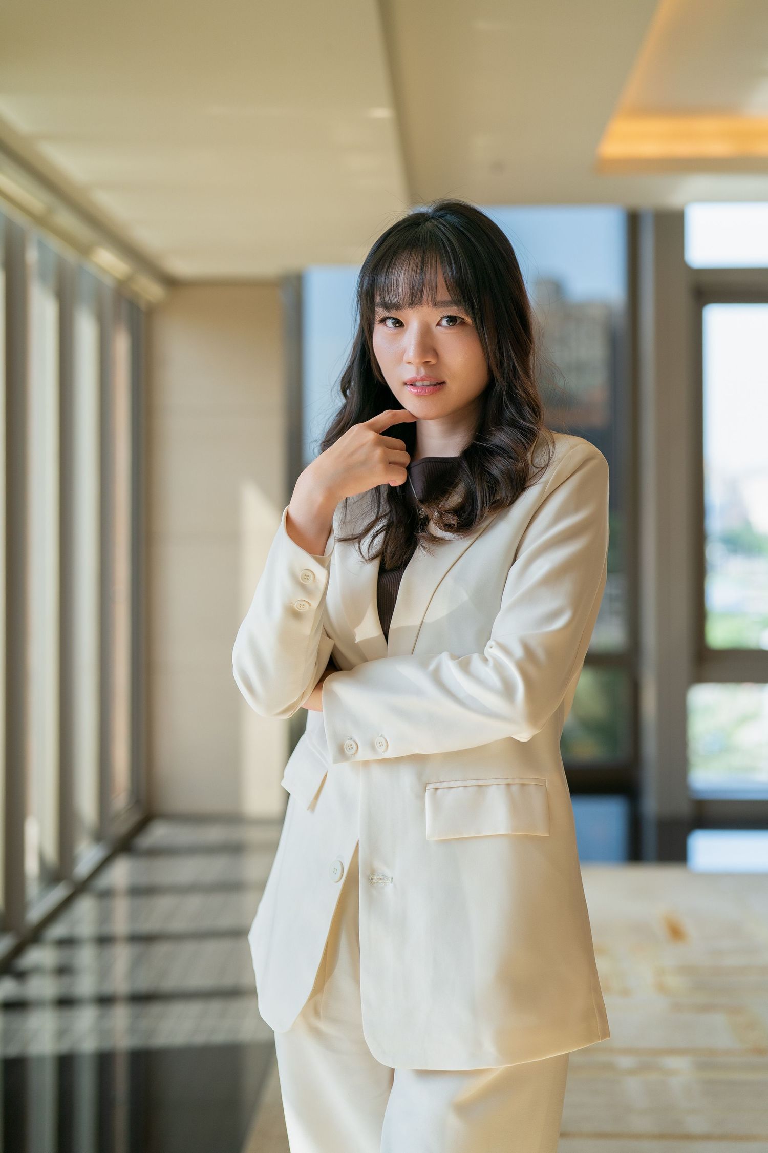 Young Asian businesswoman in cream suit posing confidently in modern office hallway with natural light.