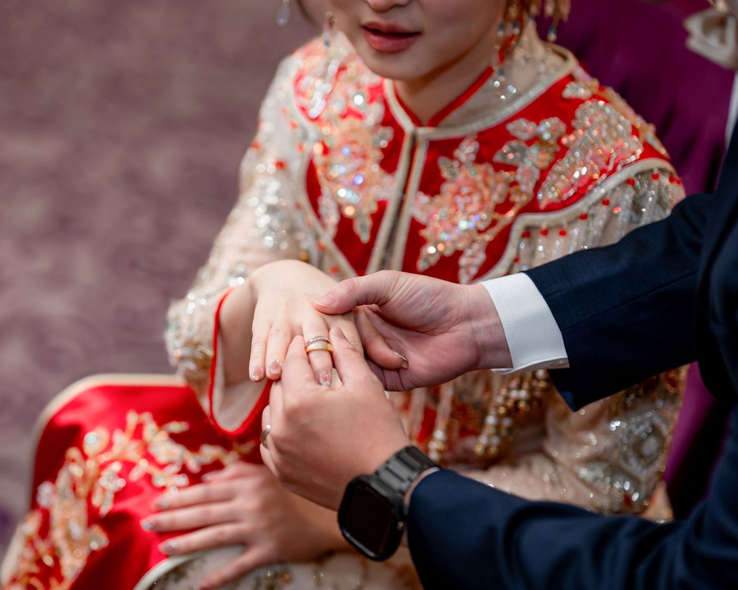 Groom in a navy suit places a wedding ring on a bride wearing a red and gold embroidered traditional Chinese gown.