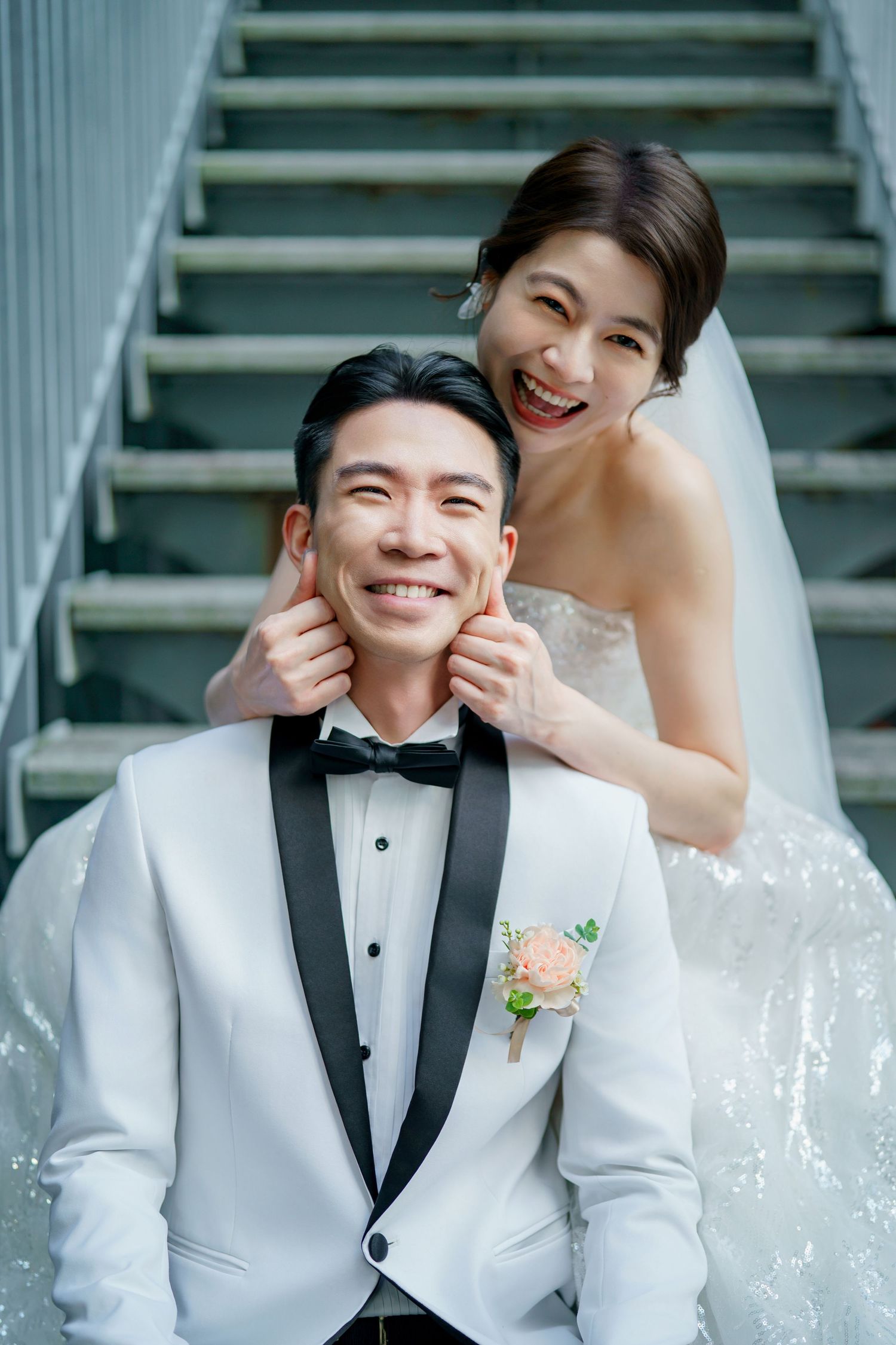 Joyful wedding couple sharing a playful moment on stairs with bride in white dress and groom in white tuxedo.