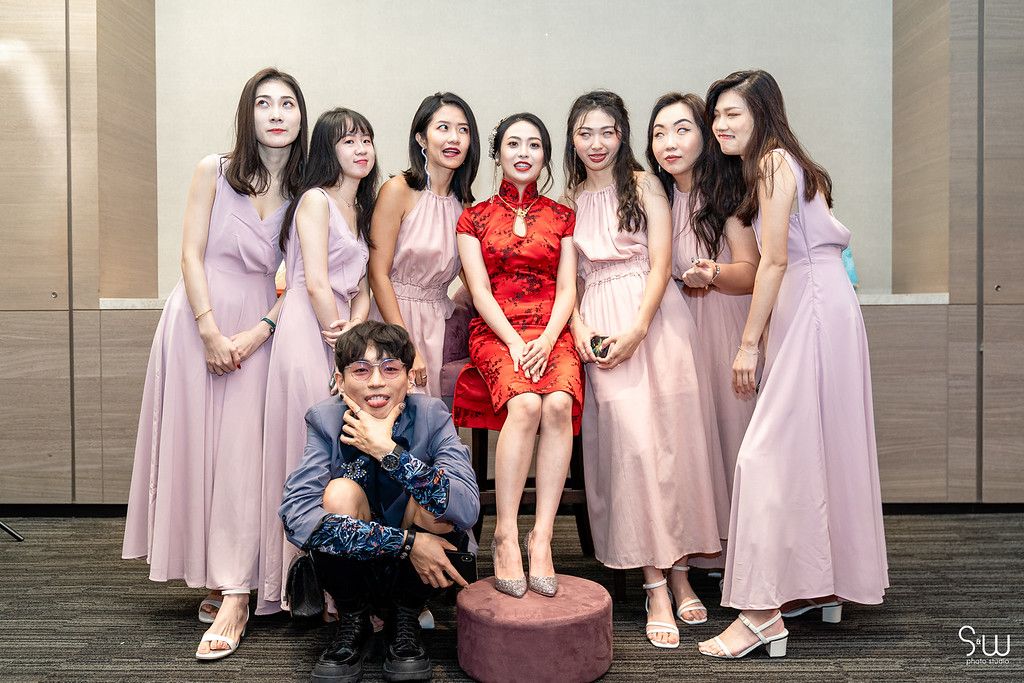 A wedding party poses in pink dresses with one person wearing a traditional red qipao dress.