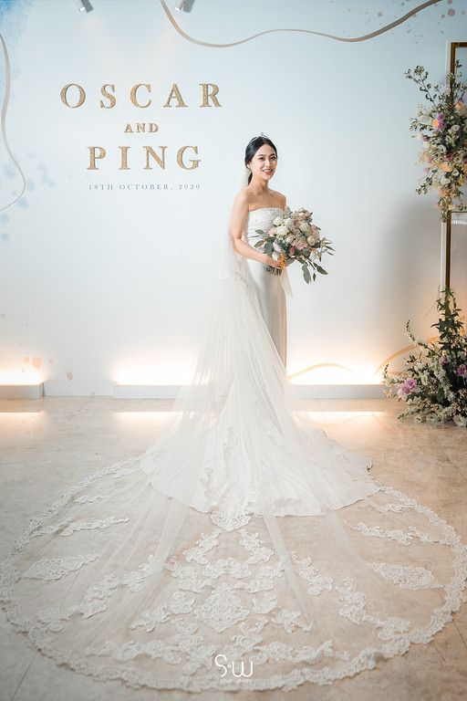 Bride in lace train wedding dress poses elegantly against wall with floral decorations.