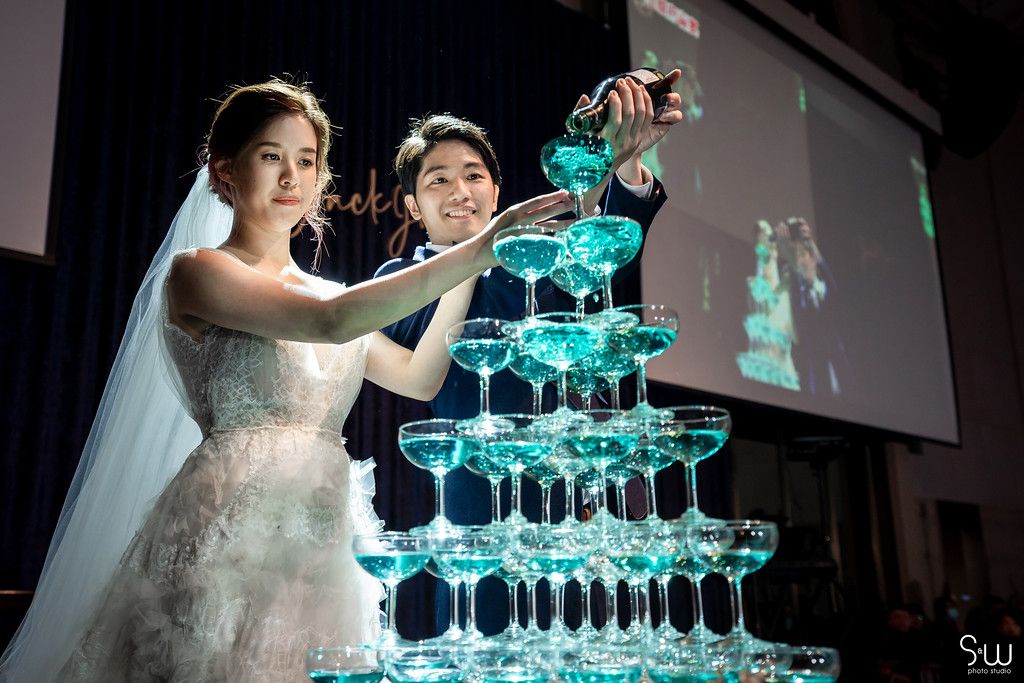 Wedding couple pours champagne into tower of glasses with blue lighting.