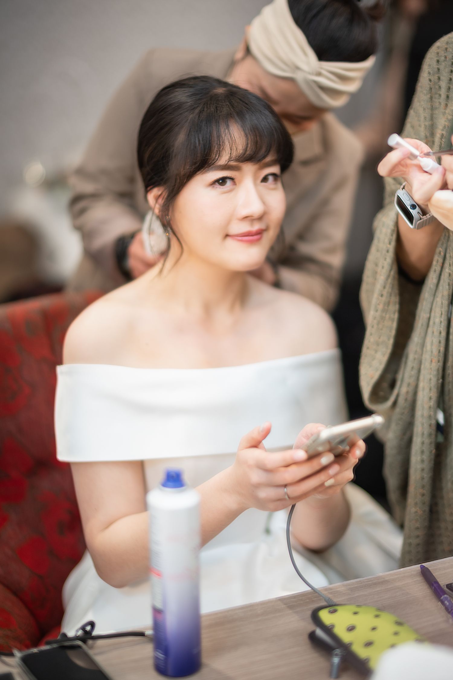Bride getting ready in off-shoulder white dress while holding phone during wedding preparations.